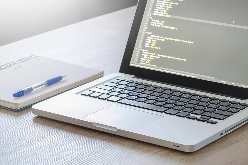 حاسوب محمول مفتوح تظهر على شاشته أسطر من كود CSS، وبجانبه دفتر وقلم أزرق