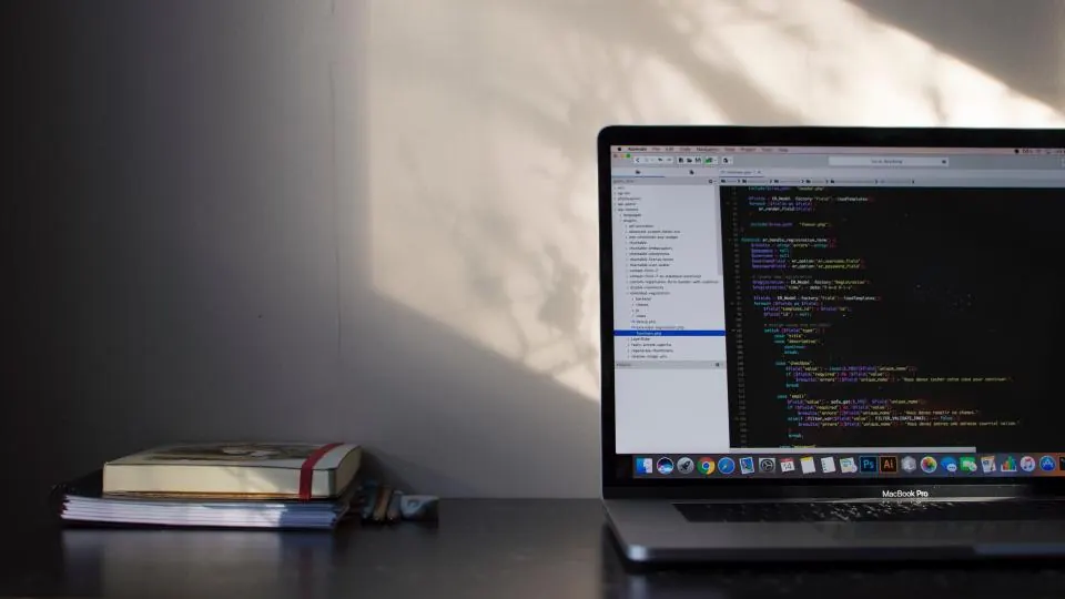 A laptop on a dark desk showing lines of code, next to a stack of books in soft daylight