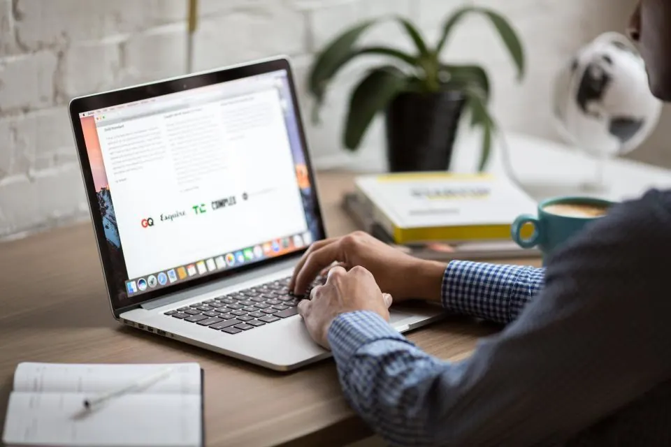 A man typing on a laptop in a bright office next to a plant, a coffee cup and a notebook